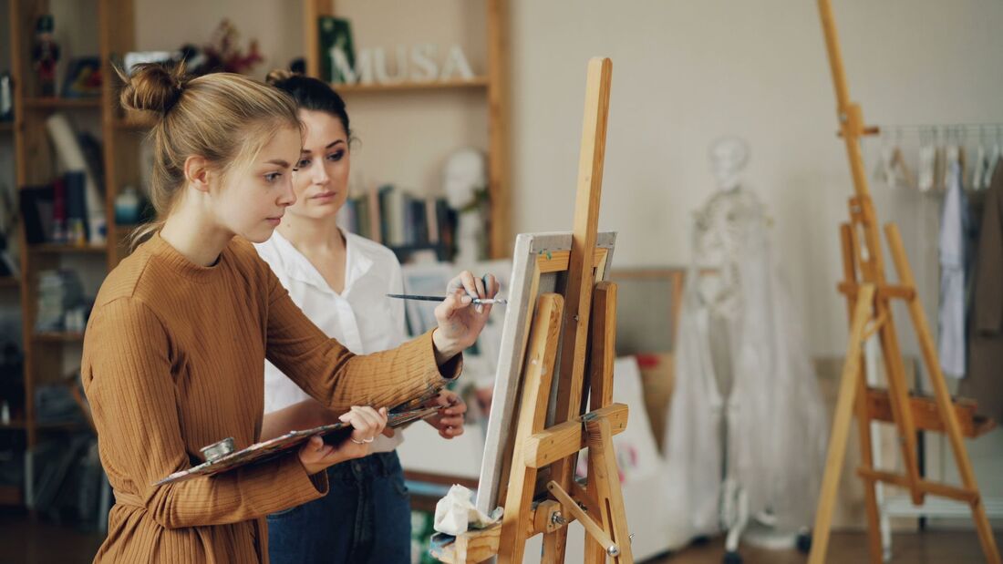 An art student painting at an easel while a tutor observes